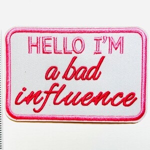 May include: A rectangular white patch with a pink border and the embroidered text "HELLO I'M a bad influence" in pink. The patch is approximately 3 inches long.