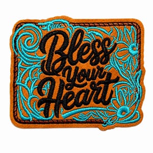May include: A rectangular embroidered patch with a tan felt base. The patch features the black stitched text "Bless your heart" in a cursive script, surrounded by intricate turquoise floral scrollwork and a dark brown stitched border.