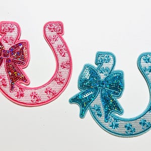 May include: Two embroidered horseshoe patches. One is pink with a sequined bow, the other is blue with a sequined bow. Both have floral patterns and white outlines. These patches are ideal for adding flair to clothing or accessories.