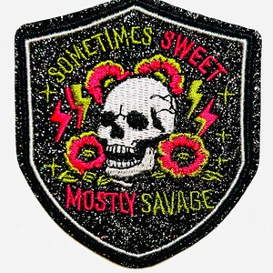 May include: A black, glittery patch in a shield shape, featuring a white skull with pink and yellow flowers, and red and yellow lightning bolts. The text reads "Sometimes Sweet, Mostly Savage."