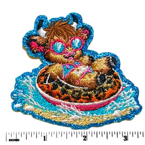 May include: Embroidered patch featuring a cartoon cow wearing sunglasses and a swimsuit, floating on an inner tube in a pool. The patch has a blue glitter border and is on a white background. The cow is brown, tan, and white.