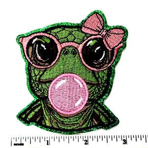 May include: Embroidered patch featuring a green turtle with pink sunglasses and a pink bow. The turtle is blowing a pink bubble. The patch has a green glitter border. The patch is approximately 3 inches wide.
