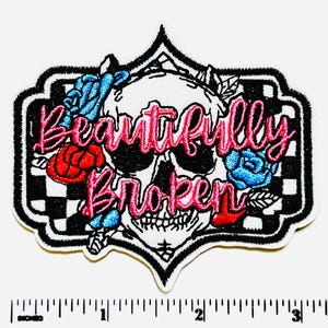 May include: Embroidered patch featuring a skull with the words "Beautifully Broken" in pink script. The skull is surrounded by red and blue roses, set against a black and white checkered background, with a black border.