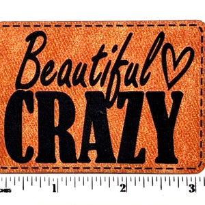 May include: A rectangular tan patch with black stitching around the edges and the black text Beautiful CRAZY with a heart symbol. A white measuring tape is positioned along the bottom edge, showing a width of approximately 3.25 inches.