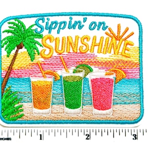 May include: A rectangular embroidered patch featuring the text Sippin' on SUNSHINE above three glasses of colorful drinks on a beach with a palm tree and sunset. A ruler at the bottom shows the patch is approximately 3.25 inches wide.