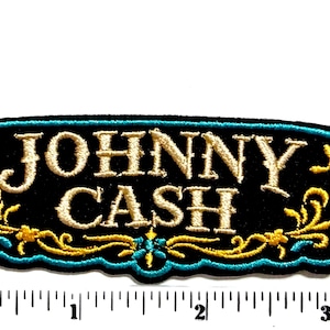 May include: A rectangular embroidered patch with a black background featuring the text JOHNNY CASH in cream-colored thread. The patch has a teal border and gold filigree scrollwork at the bottom. A ruler shows the patch is 3.5 inches wide.