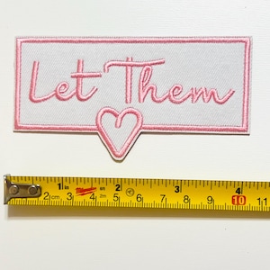 May include: A rectangular, white embroidered patch with the words "Let Them" in pink script, framed by a pink border. A pink heart shape extends from the bottom. A yellow tape measure is placed below the patch.