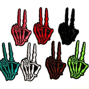 May include: A collection of seven embroidered patches in the shape of a hand making the peace sign. The patches are in various colors, including red, black, green, and white, with a skeleton hand design. The patches are on a white background.