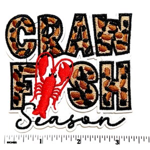 May include: Embroidered patch with the words "CRAWFISH SEASON" in black and leopard print lettering. A red crawfish is in the center. The patch is on a white background with an inch ruler at the bottom.