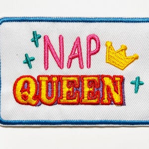 May include: Embroidered patch with a white background and a blue border. The patch features the words "NAP QUEEN" in pink and yellow, with a gold crown and teal starburst accents. The patch measures approximately 2 inches by 3 inches.