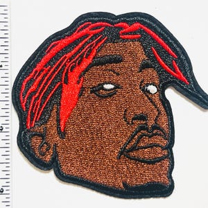 May include: Embroidered patch featuring a stylized portrait with brown skin, red hair, and a black outline. The patch is approximately 3 inches tall and 2.5 inches wide, suitable for application on clothing or accessories.