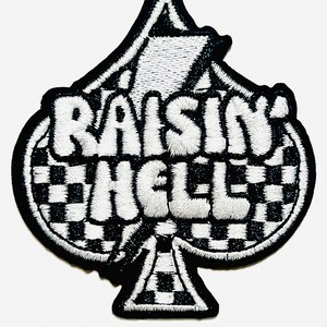 May include: Black and white embroidered patch in the shape of a spade. The patch features the words "RAISIN HELL" in white, puffy letters. The spade's background is a black and white checkered pattern.