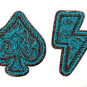 May include: Two embroidered patches: a turquoise spade and a turquoise lightning bolt. Both have a brown glitter border and turquoise filigree designs. The patches are set against a white background.