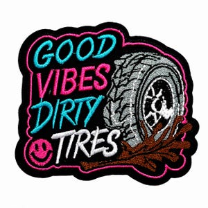 May include: An embroidered patch with a black background featuring the text GOOD VIBES in blue and pink, DIRTY in blue, and TIRES in white. A grey tire with brown mud splatter and a small pink smiley face icon are included in the design.