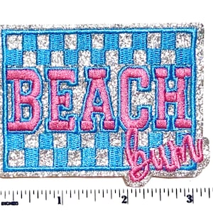 May include: Embroidered patch with a rectangular shape, featuring the words "BEACH" in pink block letters and "bum" in cursive, outlined in blue. The background has a blue and white checkered pattern, with a silver glitter border.