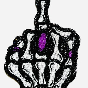 May include: Embroidered patch featuring a black and white skeleton hand giving the middle finger. The patch has a glittery black border and a purple accent in the center of the hand.