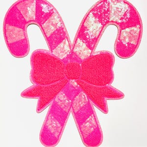 May include: A pair of pink sequined candy canes with a matching bow. The candy canes have alternating pink and white diamond patterns. The bow is centered between the candy canes, creating a festive holiday decoration.