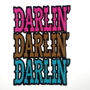 May include: Three embroidered patches with the word "DARLIN'" in a Western-style font. The patches are in pink, brown, and teal, each with a black border. The patches are stacked vertically.