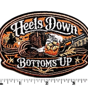 May include: Embroidered patch with the words "Heels Down Bottoms Up" in white script. The patch features a cowboy boot, a beer mug, and a rifle. The background includes a silhouette of a cowboy on a horse and a sunset.
