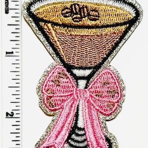 May include: Embroidered patch in the shape of a martini glass with a pink bow. The martini glass has a gold-colored drink with coffee beans. The patch is approximately 2 inches tall.