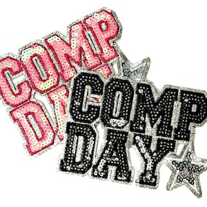May include: Two sequined patches with the words "COMP DAY". One patch is pink with silver trim, the other is black with silver trim. A silver star is next to the black patch. The patches are likely for clothing or accessories.