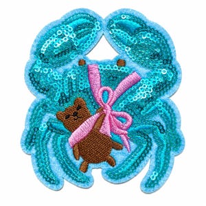 May include: A decorative patch featuring a bright turquoise crab covered in shimmering sequins. Centered on the crab is a small brown embroidered teddy bear holding a vibrant pink ribbon tied into a bow, with the entire design on a light blue felt base.