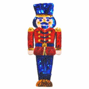 May include: A sequined nutcracker patch featuring a blue hat, a red jacket with gold buttons, and blue trousers. The design is embellished with shiny, colorful sequins throughout, creating a textured surface that depicts a soldier figure.