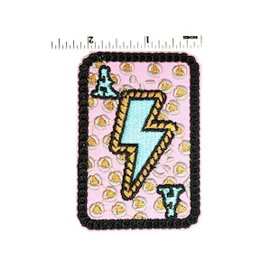 May include: A rectangular patch with a pink background featuring a light blue lightning bolt design outlined in gold. The patch has a black border and a pattern of small yellow circles. The patch is approximately 2 inches in height.