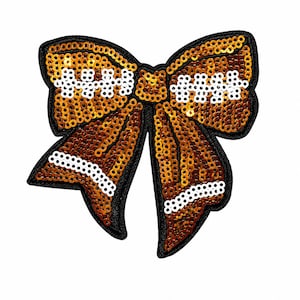 May include: A decorative bow-shaped patch covered in shimmering orange sequins with white sequin accents forming horizontal stripes. The entire shape is outlined in a thick black border, creating a distinct contrast against the bright orange and white.