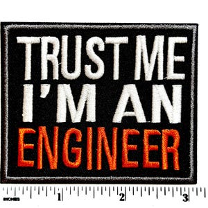 May include: Black square patch with white embroidered text that reads "TRUST ME I'M AN ENGINEER". The word "ENGINEER" is in orange embroidery. The patch has a black border and is measured in inches.