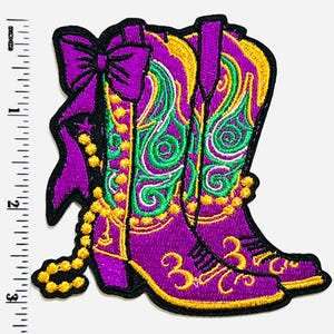 May include: Embroidered patch featuring a pair of purple cowboy boots with intricate green and gold detailing. A purple bow and beaded necklace accent the boots. The patch measures approximately 3 inches in height.