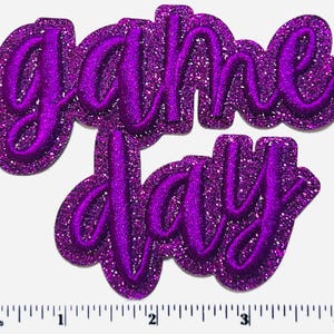 May include: A purple glitter patch with the words "Game Day" in a cursive font. The patch has a raised, textured appearance and is outlined in a darker purple. The background is white.