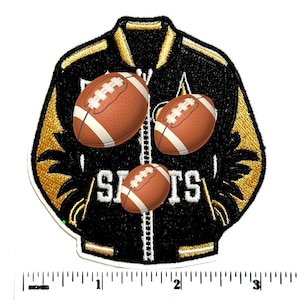May include: A black and gold varsity jacket patch featuring three brown footballs and the white text "SAINTS" across the front. A white ruler is positioned at the bottom, showing the patch width is approximately 3.5 inches.