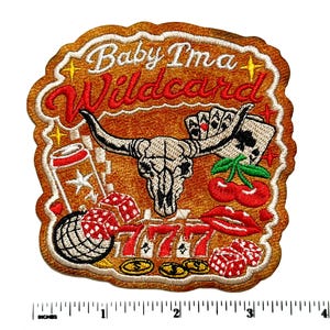 May include: An embroidered patch with a brown background featuring a steer skull, playing cards, red cherries, dice, and the text "Baby I'ma Wildcard" in white and red. A ruler at the bottom shows the patch is approximately 4 inches wide.