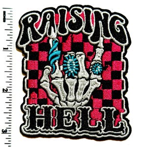 May include: Embroidered patch with the words "RAISING HELL" in black and white, featuring a skeleton hand with turquoise accents against a red and black checkered background. The patch measures approximately 3 inches in height.