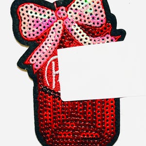 May include: A red sequined gift patch with a black border and a pink sequined bow. The patch has a white rectangular space for text. The patch measures approximately 2 inches in height.