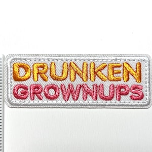 May include: Embroidered patch with the words "DRUNKEN GROWNUPS" in orange and pink lettering. The text is on a white rectangular patch with a white border. The patch is on a white surface.