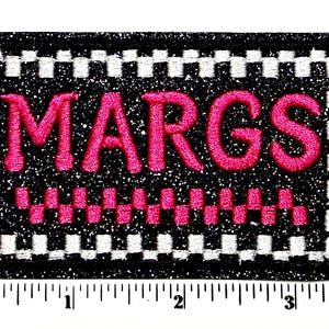 May include: A rectangular patch with the word "MARGS" embroidered in bright pink on a black glitter background. The patch is bordered with a white and black checkered pattern and measures approximately 3 inches wide.