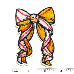 May include: An embroidered patch shaped like a bow with long, flowing tails. The design features a yellow and white pattern with red stitching that mimics the appearance of a softball. A white ruler shows the patch is approximately 3.5 inches wide.