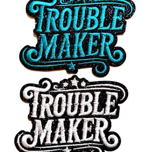 May include: Two embroidered patches with the words "TROUBLE MAKER". One patch features turquoise text on a black background with glitter accents. The other patch has white text on a black background.