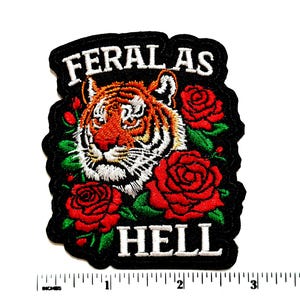 May include: An embroidered patch featuring a tiger head surrounded by red roses and green leaves on a black background. The text FERAL AS HELL is stitched in white. A ruler at the bottom shows the patch is approximately 3.5 inches wide.