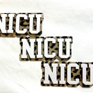 May include: Three rectangular pins with the letters "NICU" in white block letters. The background of the pins has a tan and black leopard print pattern. The pins are arranged in a diagonal line.
