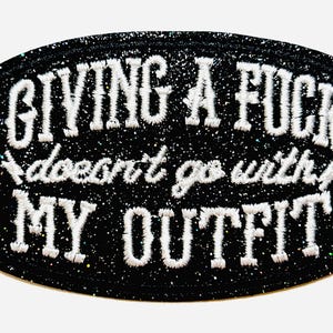 May include: Black, glitter-covered patch with white embroidered text: "GIVING A FUCK doesn't go with MY OUTFIT." The patch is approximately 2 inches in height. A humorous accessory for clothing or bags.