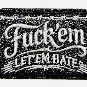 May include: Black rectangular patch with white embroidered text that reads "Fuck'em LET'EM HATE". The patch has a glittery texture and a decorative border. The patch is approximately 2 inches in height.