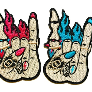 May include: Two embroidered patches featuring a hand gesture with the index and pinky fingers extended. One patch has red flames, the other has blue flames. Both have a cigarette, nail polish, and a lightning bolt.