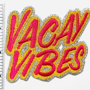 May include: Embroidered patch with the words "VACAY VIBES" in bright pink script. The text is outlined in gold and set against a silver glitter background. The patch is approximately 3 inches wide and 2.5 inches tall.