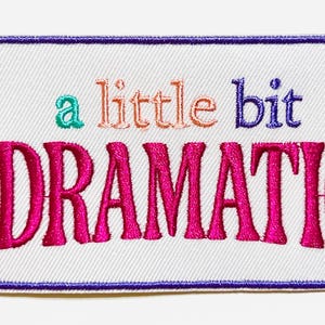 May include: Embroidered patch with the phrase "a little bit DRAMATIC" in teal, orange, purple, and hot pink. The patch has a white background and a purple border. The patch is rectangular.