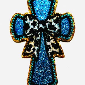 May include: A decorative cross-shaped applique with a blue glitter design and gold trim. The center features a black and white leopard print pattern. The cross measures approximately 2 inches in length.
