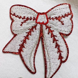 May include: A white bow with red glitter trim. The bow is embroidered with a red design.