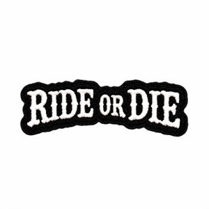 May include: A black embroidered patch with a white stitched border and the text RIDE OR DIE in white capital letters. The patch has a curved, irregular shape that follows the contour of the bold, serif-style lettering across the center.